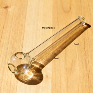 Clear glass hand pipe labeled with mouthpiece and bowl resting diagonally on light wooden surface