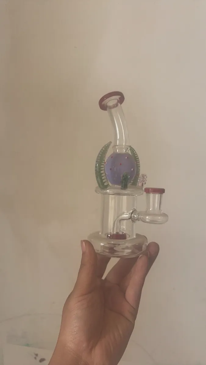 Hand holding decorative glass bong with purple sphere, green leaf ornaments and red caps