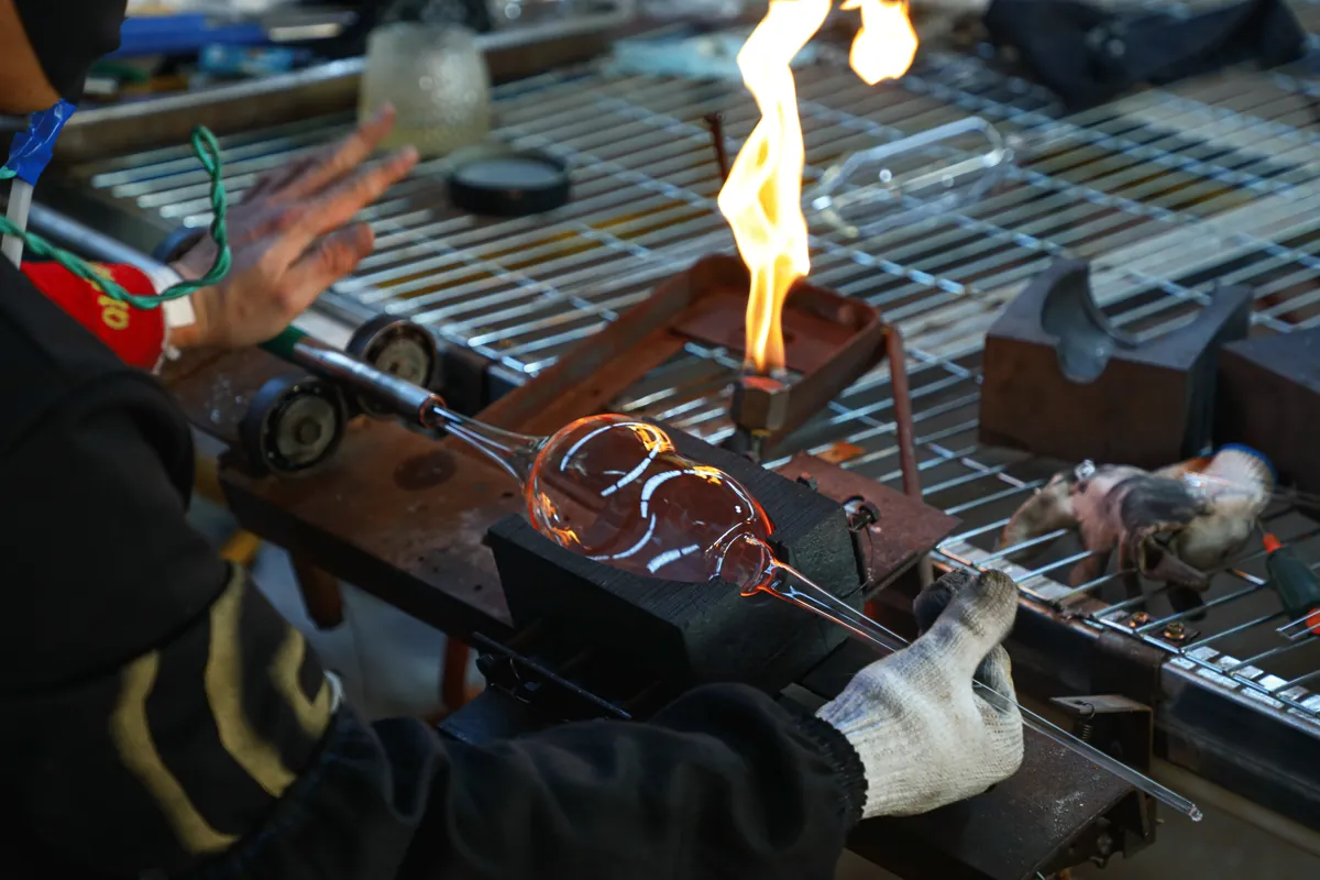 Glass artisan shaping glowing molten glass on blowpipe with torch flame
