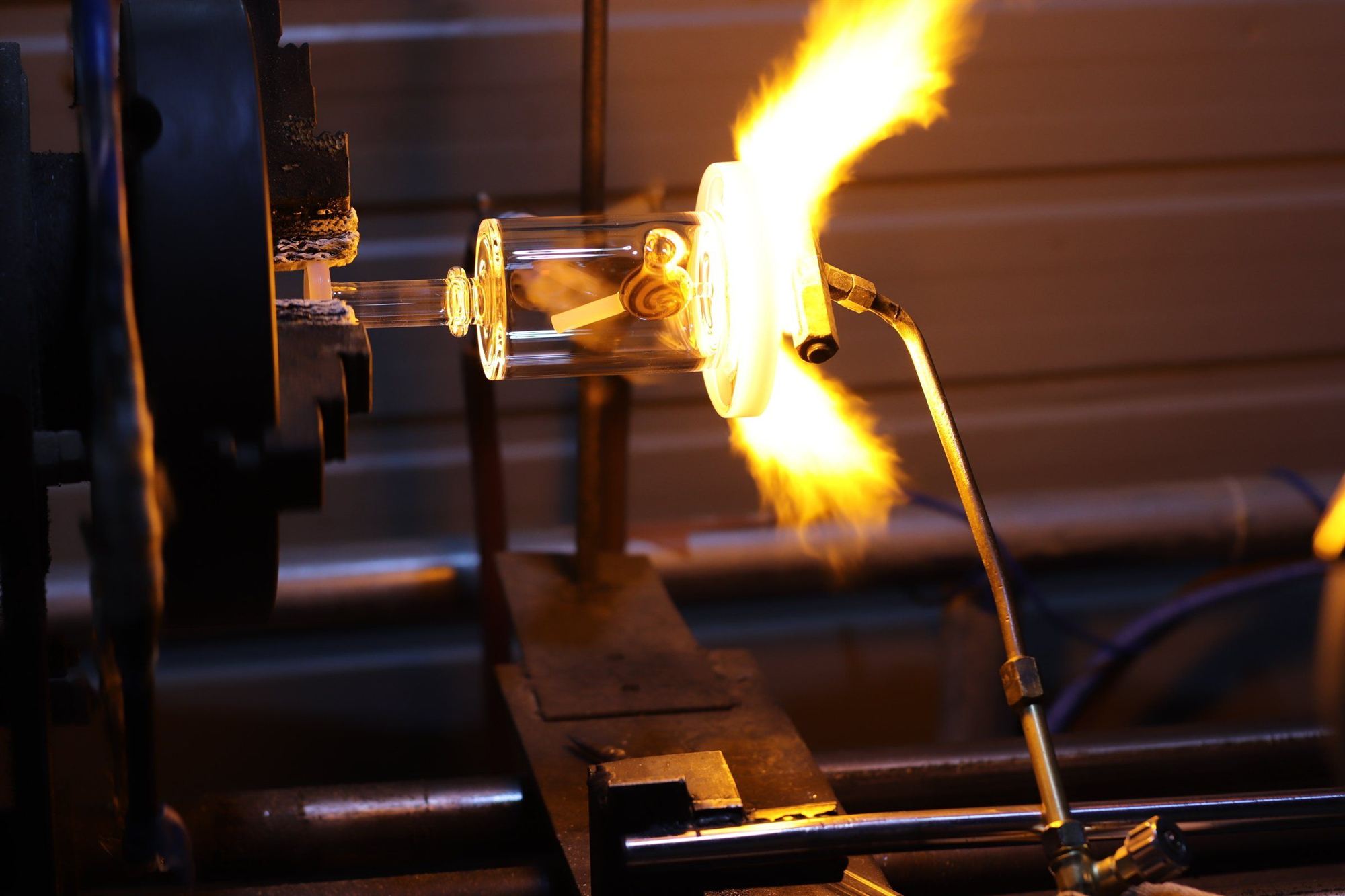 Glassblower heating a clear glass tube with bright torch flame on a metal lathe fixture