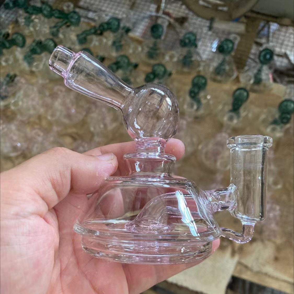 Hand held pink tinted glass dab rig with percolator chamber in workshop with other glass pieces blurred